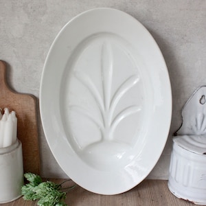 May include: A large, white, oval serving platter with a raised botanical design in the center. The platter is surrounded by a wooden cutting board, a ceramic container holding white candles, and a white wall-mounted salt cellar.