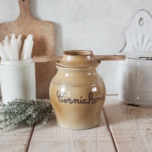 Puede incluir: Un tarro de cerámica beige con tapa y cuchara de madera, etiquetado "Cornichons" en cursiva. El tarro está sobre una superficie de madera, con un recipiente de cerámica blanca con velas y una tabla de cortar de madera en el fondo.