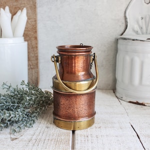 Puede incluir: Una pequeña lata de leche de cobre con un asa y un borde de latón. La lata tiene una textura martillada y está sobre una superficie de madera blanca.