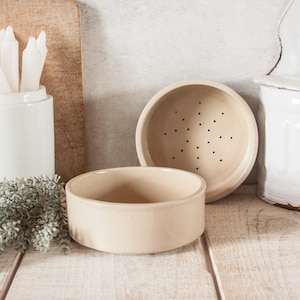 May include: Two beige ceramic bowls, one with drainage holes, are on a light-coloured wooden surface. The bowls are round and low. A white ceramic container with utensils and a wooden chopping board are in the background.