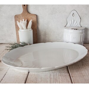 May include: A large, white, oval serving platter with a scalloped edge. The platter is on a wooden surface, with a wooden cutting board, a white ceramic container with candles, and a white wall-mounted container in the background.