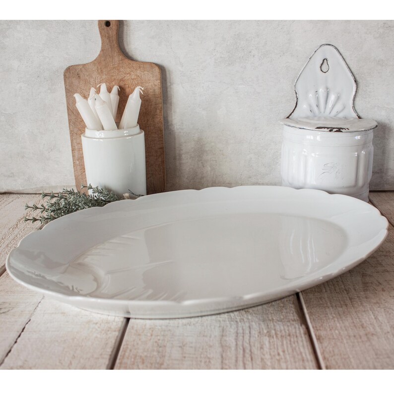 May include: A large, white, oval serving platter with a scalloped edge. The platter is on a wooden surface, with a wooden cutting board, a white ceramic container with candles, and a white wall-mounted container in the background.