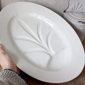 May include: An oval, white ceramic platter with a raised leaf design in the center. The platter is held by a person. The background includes a white container and glassware.