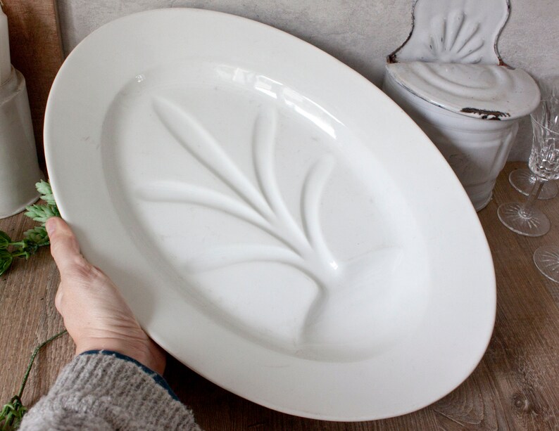 May include: An oval, white ceramic platter with a raised leaf design in the center. The platter is held by a person. The background includes a white container and glassware.