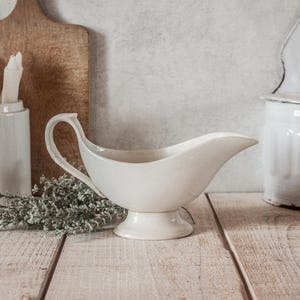 Può includere: Una salsiera in ceramica bianca con manico curvo e base a piedistallo. La salsiera è appoggiata su una superficie di legno chiaro, con un tagliere di legno e altri oggetti in ceramica bianca sullo sfondo. Un rametto di verde è posto vicino alla salsiera.