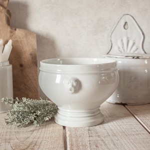 Puede incluir: Un cuenco de cerámica blanca con base de pedestal y un pequeño detalle de cabeza de león. El cuenco forma parte de un juego de utensilios de cocina de cerámica blanca, que incluye un tarro con tapa. El cuenco está sobre una superficie de madera.