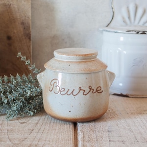 May include: A cream-coloured ceramic butter crock with a speckled finish and a matching lid. The word Beurre is written in brown cursive script on the front. The container features two small side handles and a base with a brown gradient.