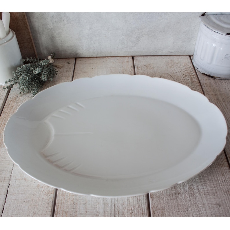 May include: A large, white, oval serving platter with a scalloped edge. The platter has a smooth surface with subtle embossed details. The design is simple and elegant, suitable for serving a variety of dishes. The platter is made of ceramic.