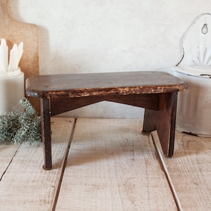 Può includere: Un piccolo sgabello in legno antico con finitura marrone scuro. Lo sgabello ha un piano rettangolare e quattro gambe. Il legno mostra segni di usura, aggiungendo al suo fascino vintage. Lo sgabello è probabilmente utilizzato per scopi decorativi.
