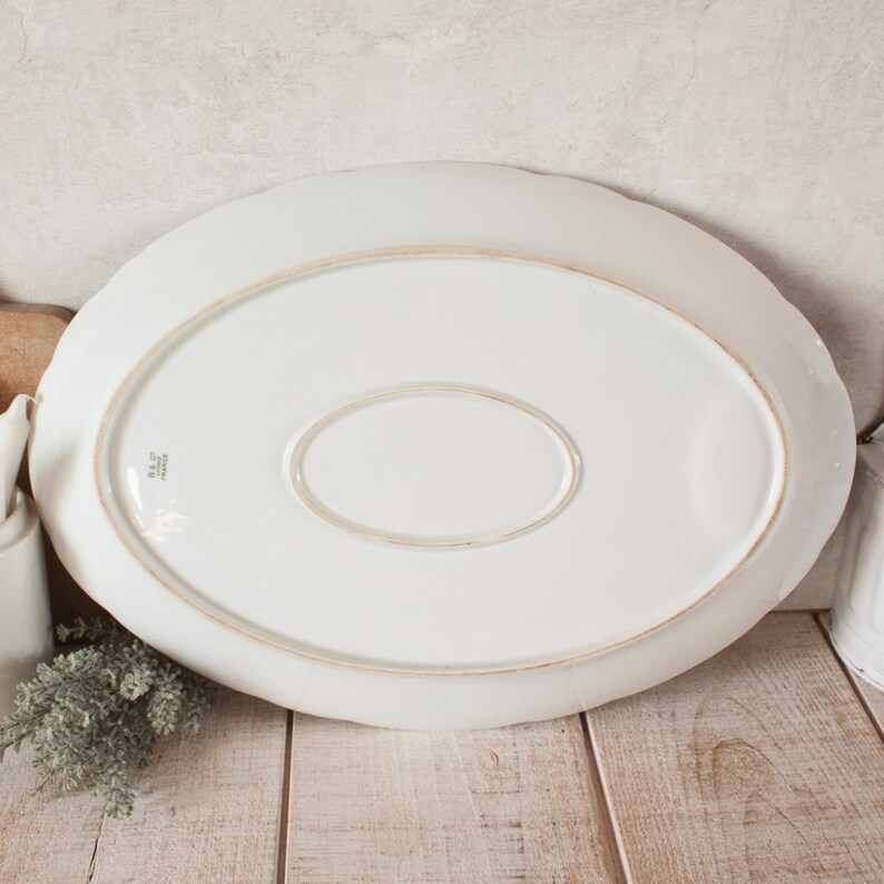 May include: An oval, white ceramic serving platter with a scalloped edge. The underside features a recessed oval and a manufacturer's mark. The platter rests on a light-colored wooden surface.