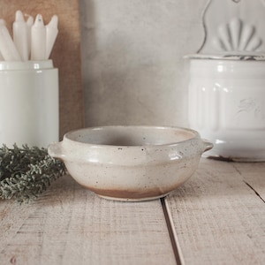 May include: A speckled, off-white ceramic bowl with two small handles. The bowl has a brown rim and sits on a wooden surface. Other items in the background include a white container with candles and a white canister.