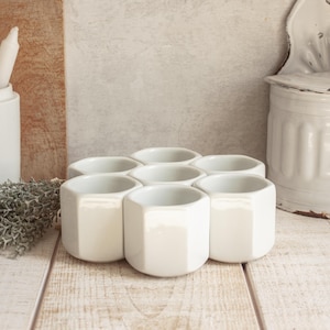 May include: A set of six white ceramic hexagonal cups arranged in a honeycomb pattern. The cups are all the same size and shape, and they are made of a smooth, glossy ceramic.