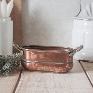 Peut inclure: Un pot en cuivre rectangulaire avec deux poignées décoratives. Le récipient présente une texture martelée et une riche patine vieillie. Le pot est présenté sur une surface en bois rustique, suggérant une esthétique de ferme ou d'antiquité.