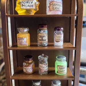 May include: A miniature brown shelf displays small glass jars with various contents and labels. The jars contain substances labeled "Honey Meade", "Mermaid Scales", "Glow Jelly", "Dragon Scales", "Frost Crystals", "Liquid Gold", "Mermaid Tea", "Glow Juice", "Worm Wood", and "Tree Tears".
