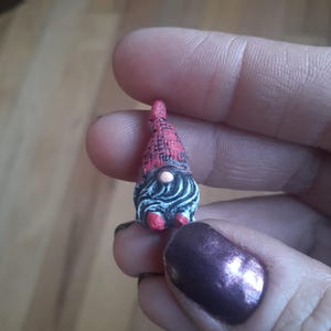 May include: A miniature gnome figurine with a red hat and shoes, and a black and white swirled beard. The gnome has a white face and a small, round nose. The figurine is small enough to be held between fingers.