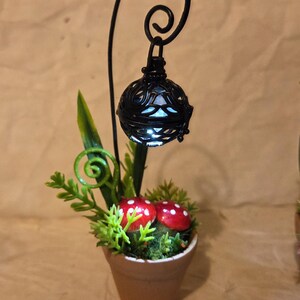 May include: A miniature fairy garden scene featuring a small terracotta pot with red mushroom accents and greenery. A black metal lantern hangs above, casting a soft glow. The whimsical design evokes a fantasy theme.