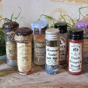 May include: A collection of small glass bottles with black or colored screw-top lids, each featuring a vintage-style paper label with text such as Essence of Frog, Mermaid Scales with Kelp, Liquid Dragon's Breath, Witch's Brew, and Swamp Water.