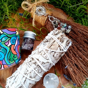 May include: A set of spiritual cleansing items including a white sage smudge stick, a small jar of clear crystals, a colorful painted terracotta pot, and a brown broom with a silver charm and crystals.