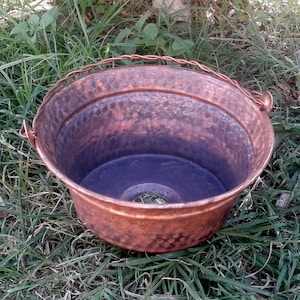 May include: A hammered copper planter with a wire handle. The planter is sitting on a bed of green grass.