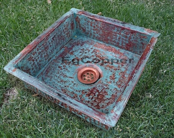 May include: A square copper sink with a hammered finish and a patina of turquoise and copper. The sink has a drain in the center.