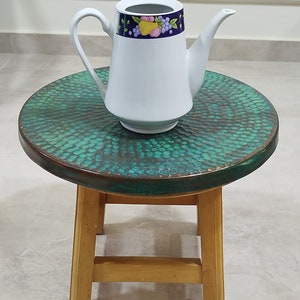 May include: A white ceramic teapot with a blue and purple floral design sits on a green and brown round stool with wooden legs.