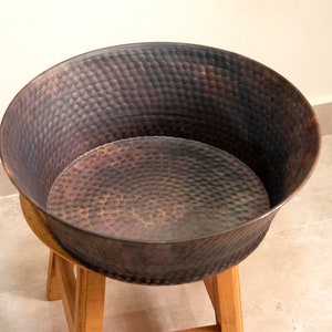 May include: A large, round, hammered copper planter sits on a wooden stand. The planter has a dark brown patina and is ideal for displaying plants or flowers.