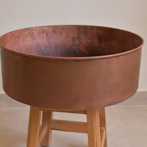 May include: A large copper planter with a rounded shape sits on a wooden stand. The planter has a slightly distressed look, giving it a vintage feel.