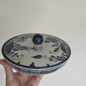 May include: A round, lidded ceramic dish with a blue and white floral design. The lid has a small, round knob. The dish is held in a hand against a plain white background. The dish has a diameter of approximately 20 cm.