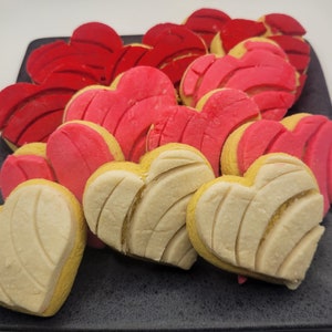 May include: A collection of heart-shaped concha pastries in shades of red, pink, and cream, arranged on a dark gray plate. The pastries have a textured, shell-like design on top, with a yellow base. A delightful treat.