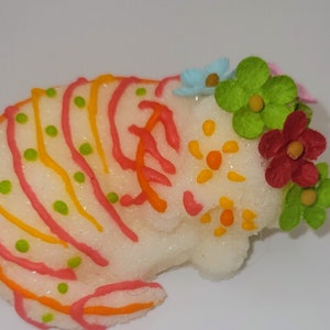 May include: A sugar skull-shaped confection, white with colorful icing details. The design includes orange, pink, and yellow lines, green dots, and a floral arrangement of red, green, and blue flowers. A traditional treat.