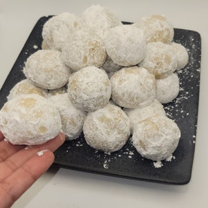 Könnte beinhalten: Ein Teller mit Schneeball-Keksen, einer Art Shortbread-Keks, der in Puderzucker gewälzt wird, auf einem schwarzen Teller.