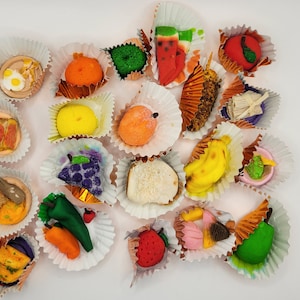 May include: Assortment of colorful, handmade fondant candies in the shape of various fruits and vegetables. The candies are displayed in individual paper cups.