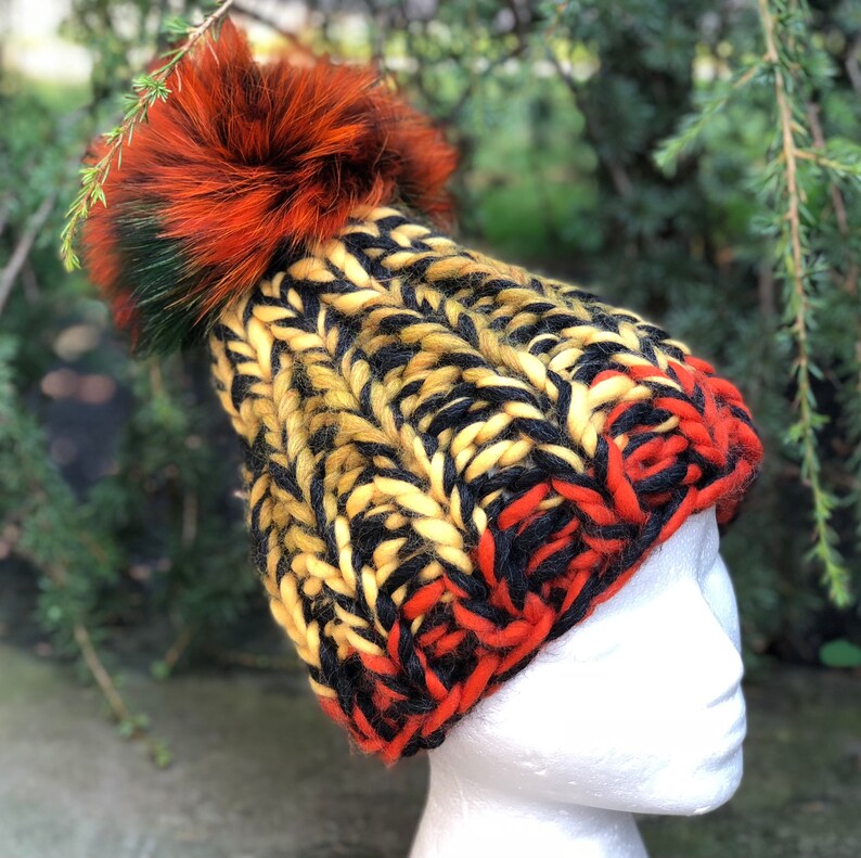 May include: A yellow, black, and red knitted beanie with a fluffy orange pom pom. The beanie is made with a chunky yarn and has a unique color pattern.