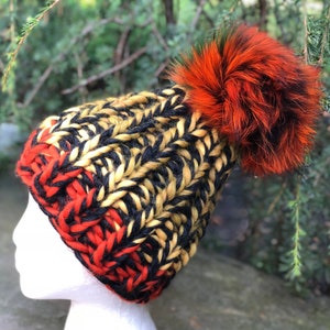 May include: A black, yellow, and red knitted beanie with a large orange and black fur pom pom.