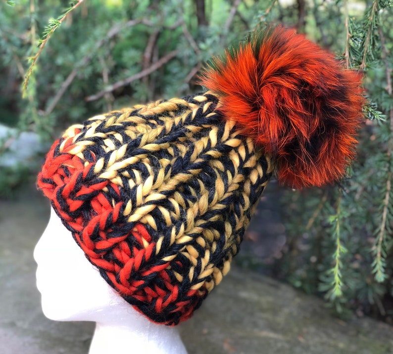 May include: A black, yellow, and red knitted beanie with a large orange and black fur pom pom.