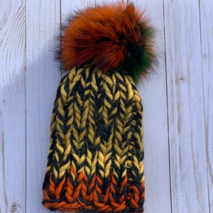 May include: A black, yellow, and orange knit beanie with a large, fluffy pom pom made of black, orange, and green fur.