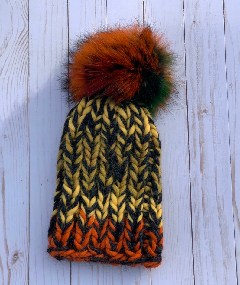 May include: A black, yellow, and orange knit beanie with a large, fluffy pom pom made of black, orange, and green fur.