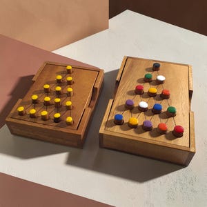 Op de afbeelding: Twee houten peg solitaire spellen. Eén spel heeft gele pinnen, de andere heeft pinnen in verschillende kleuren. De spellen zitten in houten kisten met een scharnierend deksel. De spellen staan op een wit en bruin oppervlak.