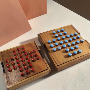 May include: Two square wooden peg solitaire game boards. One board features red cylindrical pegs arranged in a star pattern, while the other board features light blue cylindrical pegs arranged in a similar star pattern on the wooden surface.