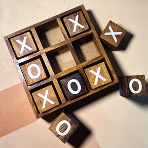 Op de afbeelding: Houten tic-tac-toe spel met een donkerbruin raster en individuele blokken. De blokken hebben witte X'en en O's. Het spel staat op een neutrale achtergrond met diagonale kleurblokken.