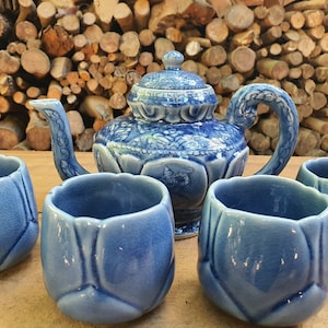 Może przedstawiać: Niebieski ceramiczny zestaw do herbaty z imbrykiem i czterema pasującymi filiżankami. Imbryk ma szczegółowy kwiatowy wzór i zakrzywiony uchwyt. Filiżanki mają kształt tulipana. Zestaw jest prezentowany na drewnianej powierzchni.