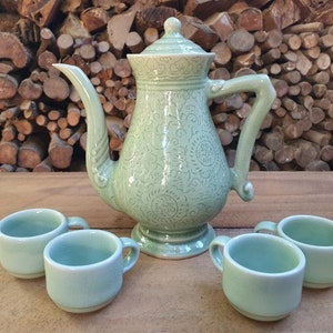 May include: A celadon green ceramic tea set with a teapot and four small cups. The teapot has a lid, a curved spout, and a handle. The set is decorated with a floral pattern and sits on a wooden surface.