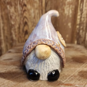 May include: A ceramic gnome figurine with a purple and white hat, a brown beard, and black eyes. The gnome is sitting on a wooden surface.