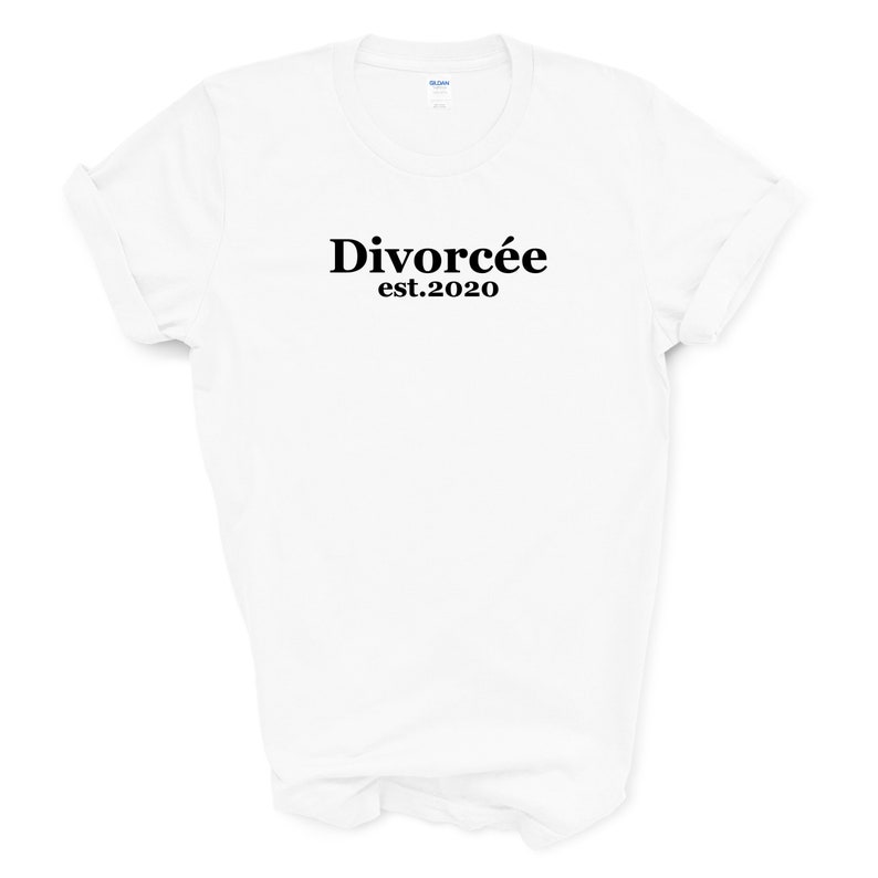 May include: White t-shirt with the word "Divorc&eacute;e" in a bold, black font, with "est.2020" below it. The shirt has rolled-up sleeves and a crew neck. The background is plain white.