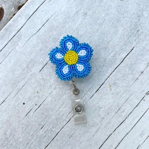 Könnte beinhalten: Ein Ausweishalter mit einer fünfblättrigen Blume aus kleinen Perlen. Die Blume hat eine leuchtend gelbe Mitte, die von weißen Perlen umgeben ist, während die äußeren Blütenblätter in Blautönen gehalten sind. Ein durchsichtiges Kunststoffband ist befestigt.