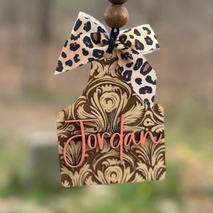 May include: A brown and tan air freshener with a floral design and the name "Jordan" in pink. It has a leopard print bow and two wooden beads at the top, hanging from a black string.