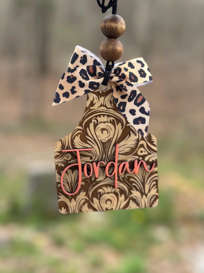 May include: A brown and tan air freshener with a floral design and the name "Jordan" in pink. It has a leopard print bow and two wooden beads at the top, hanging from a black string.