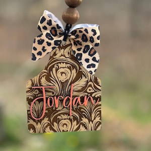 May include: A brown and gold patterned tag with the name "Jordan" in pink script, topped with a leopard print bow and wooden beads. The tag is designed to hang.