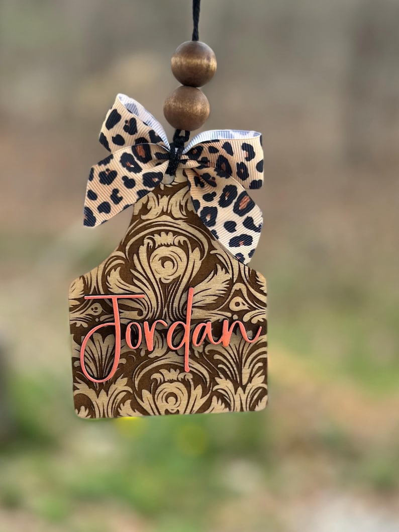 May include: A brown and gold patterned tag with the name "Jordan" in pink script, topped with a leopard print bow and wooden beads. The tag is designed to hang.