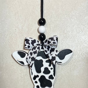 Puede incluir: Adorno con forma de vaca con dise&ntilde;o de manchas blancas y negras. Tiene una cuerda negra para colgar, con tres cuentas en blanco y negro. Un lazo con estampado blanco y negro est&aacute; atado en la parte superior de la cabeza de la vaca.